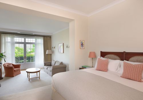 spacious hotel suite at Park Hotel Kenmare, features a neutral-toned bedroom area with a wooden headboard, pink accent pillows, and an adjacent sunken sitting room complete with a sofa, armchair, and French doors opening to a garden view.