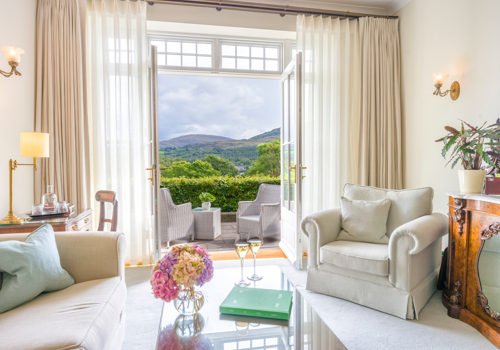 a luxurious sitting room suite at the Park Hotel Kenmare in County Kerry, Ireland, featuring cream-colored upholstered armchairs, and large French doors, open up to a private terrace overlooking Kenmare Bay and the Caha Mountains