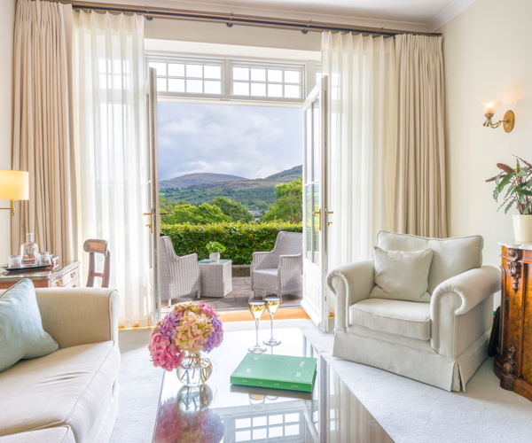 a luxurious sitting room suite at the Park Hotel Kenmare in County Kerry, Ireland, featuring cream-colored upholstered armchairs, and large French doors, open up to a private terrace overlooking Kenmare Bay and the Caha Mountains