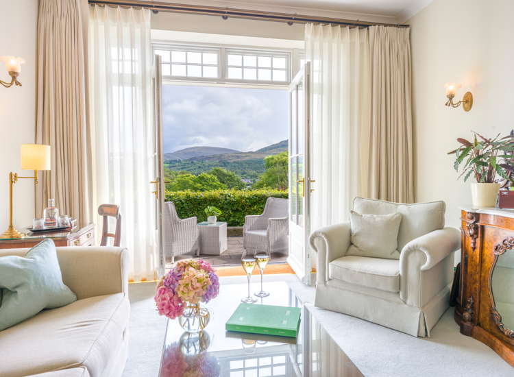 a luxurious sitting room suite at the Park Hotel Kenmare in County Kerry, Ireland, featuring cream-colored upholstered armchairs, and large French doors, open up to a private terrace overlooking Kenmare Bay and the Caha Mountains