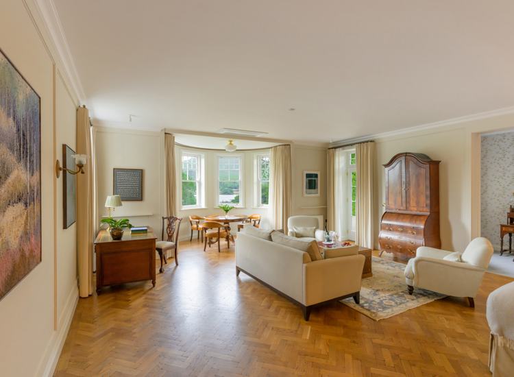 spacious Park Kenmare room features traditional furniture, herringbone hardwood flooring, and a large bay window dining nook in the background.
