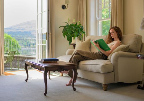 A woman reading a book on a beige sofa in a room with open doors leading to a balcony at Park Hotel Kenmare
