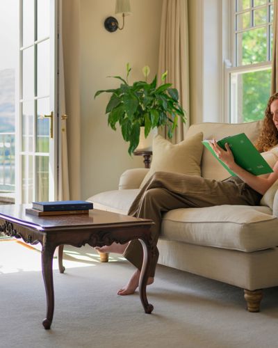 A woman reading a book on a beige sofa in a room with open doors leading to a balcony at Park Hotel Kenmare