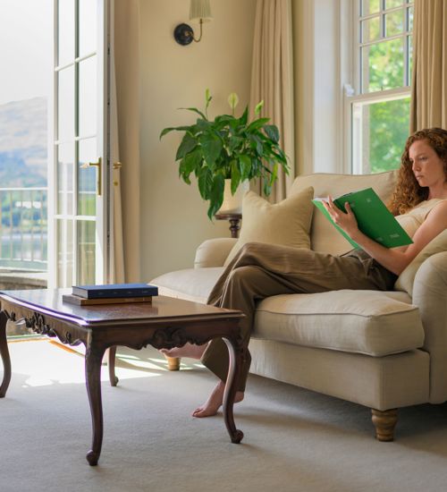 A woman reading a book on a beige sofa in a room with open doors leading to a balcony at Park Hotel Kenmare