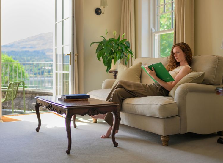 A woman reading a book on a beige sofa in a room with open doors leading to a balcony at Park Hotel Kenmare