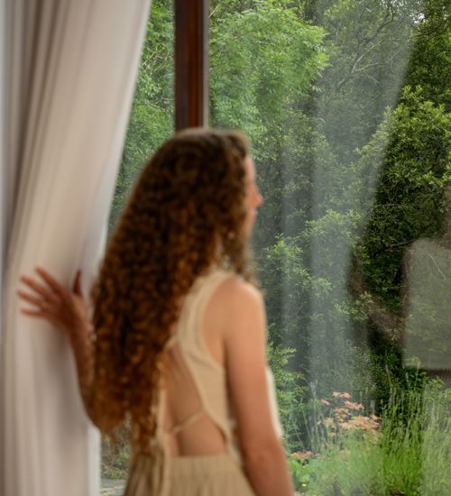 a person with curly hair looking out a large window into a lush green forest.