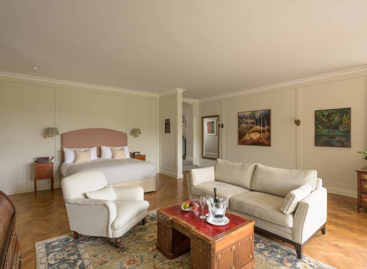 a spacious, elegantly decorated hotel suite or luxury bedroom featuring a large bed with a pink headboard, a comfortable seating area, and classic wood parquet flooring.